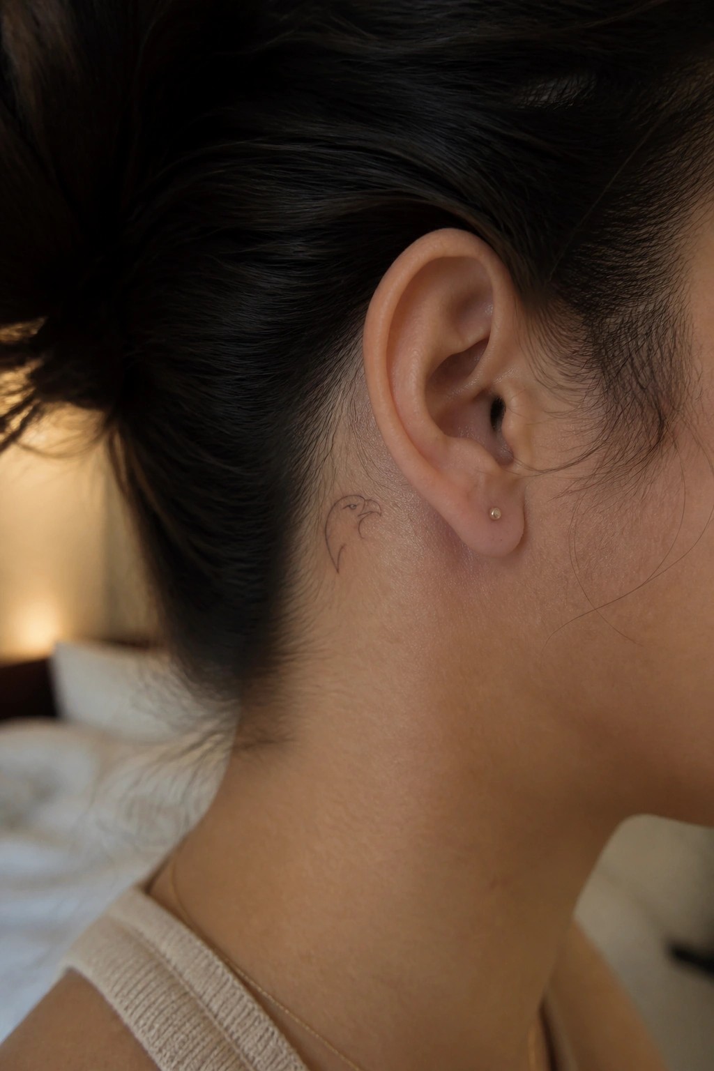 Neck with small bird tattoo behind ear, gold stud earring, dark bun, beige top.