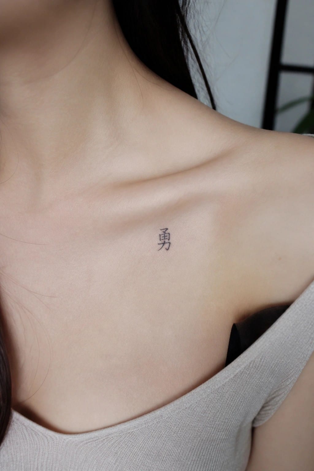 Small black Chinese character tattoo placed on the collarbone of a woman.