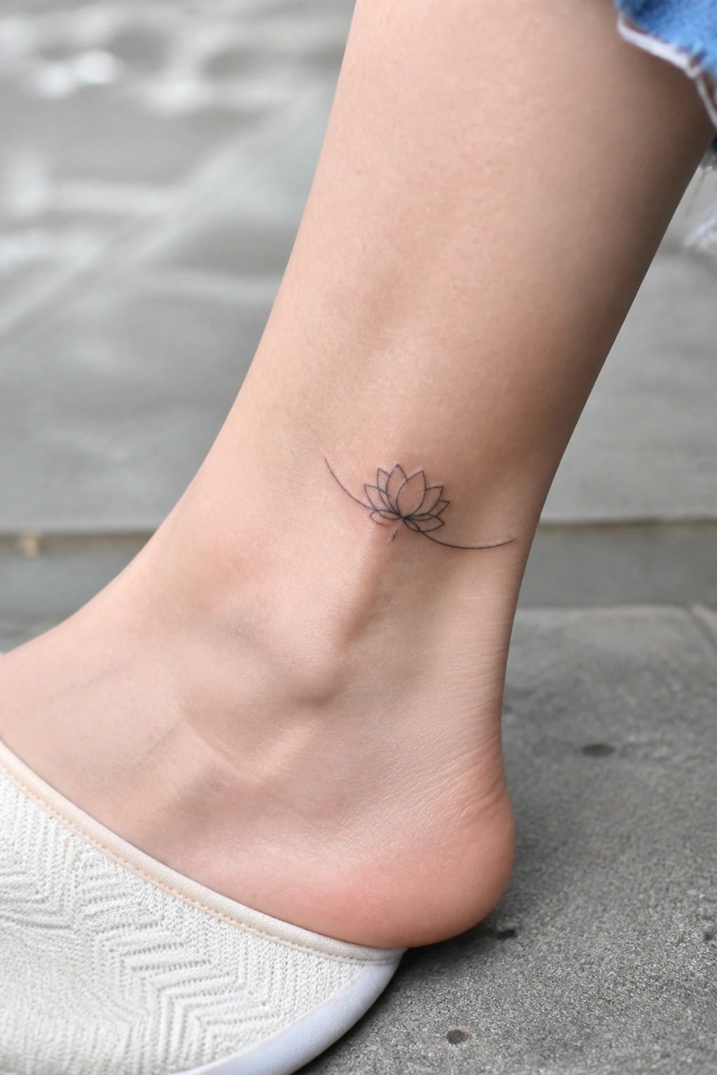 Lotus flower line tattoo on ankle above white patterned slipper on pavement