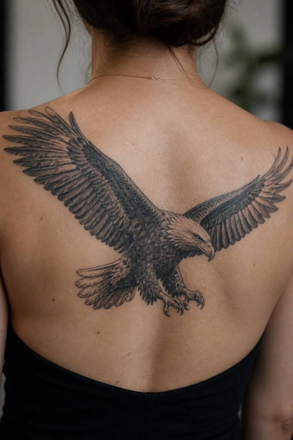 Realistic black-and-gray eagle tattoo across a woman's back with gold necklace.