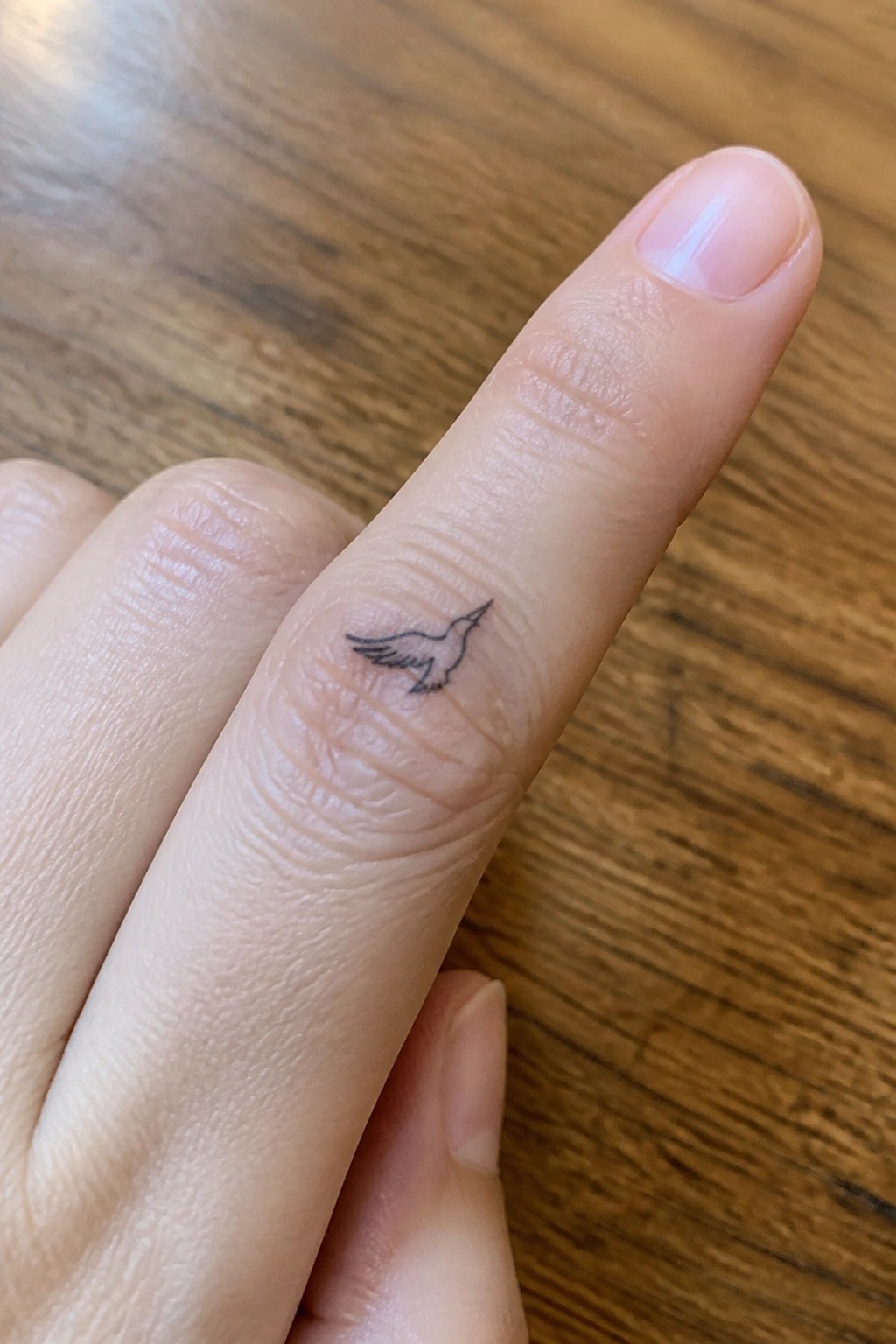 Index finger with small black bird tattoo on wooden surface.