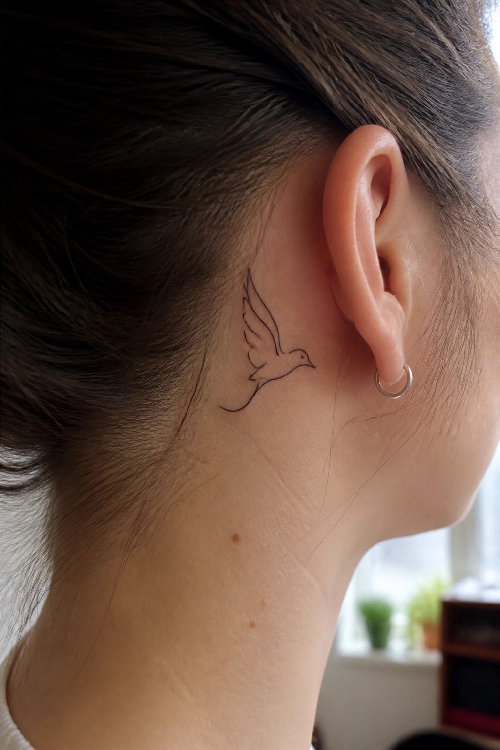 Small black bird tattoo behind ear on neck with silver hoop earring.