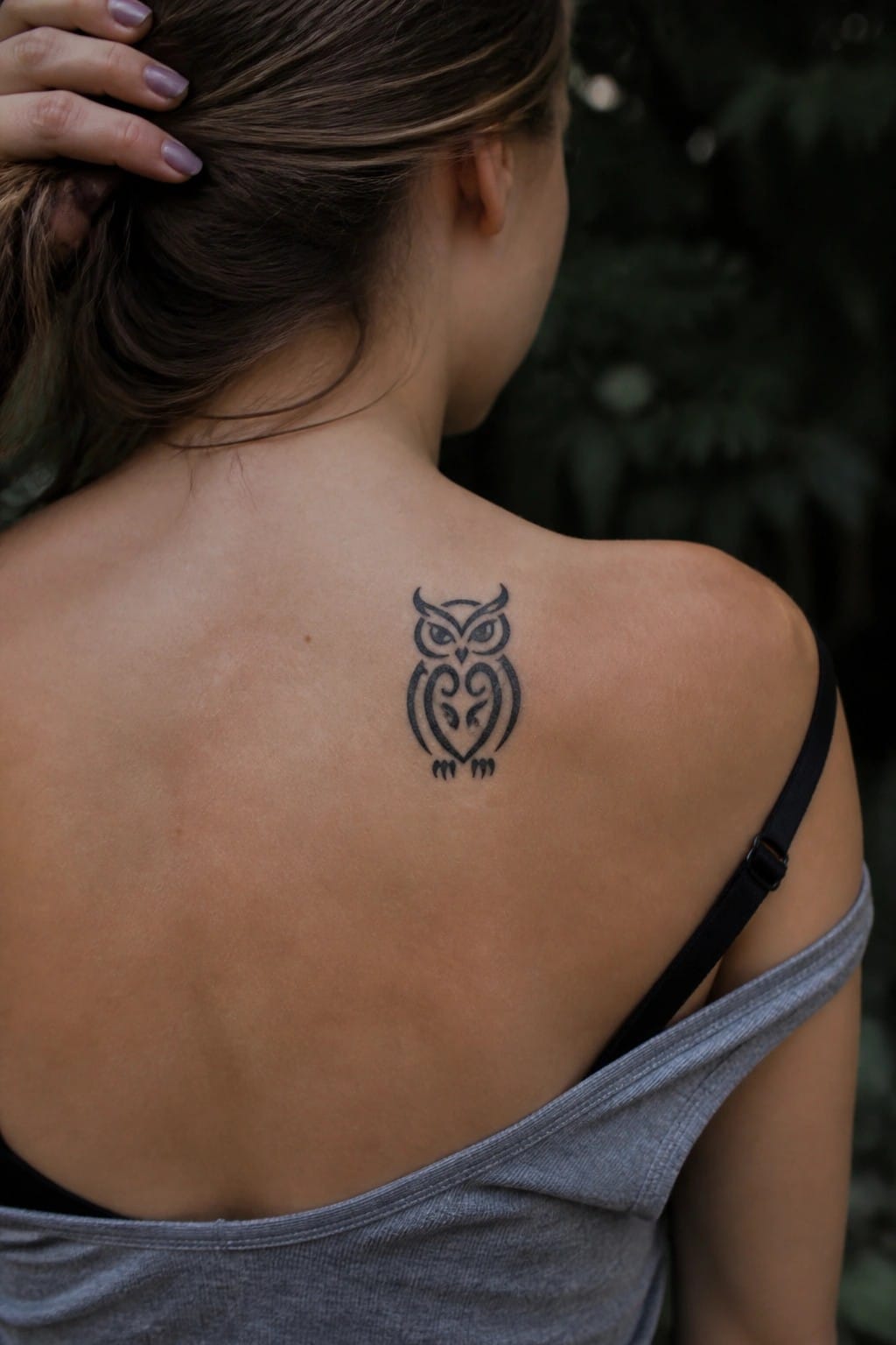 Woman's upper back with owl tattoo, brown ponytail, gray top pulled down, green background