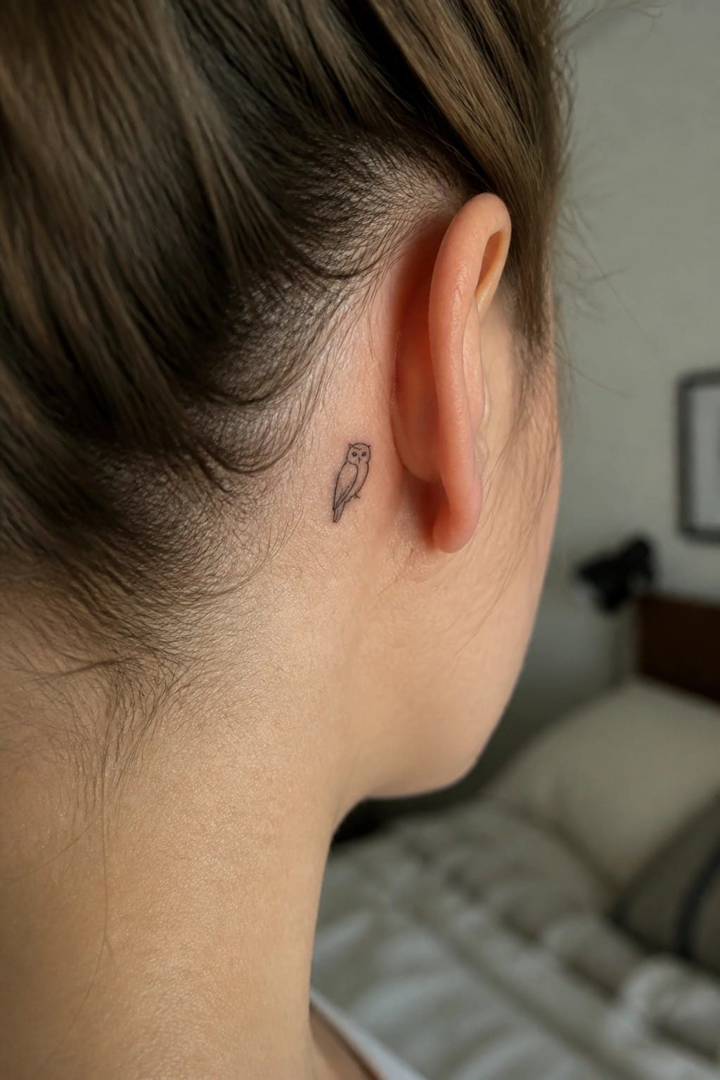 Small minimalist line drawing of an owl tattoo placed directly behind the ear on the side of the head.
