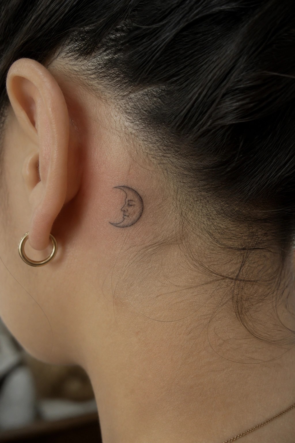 Crescent moon face tattoo behind ear with gold hoop earring and dark hair