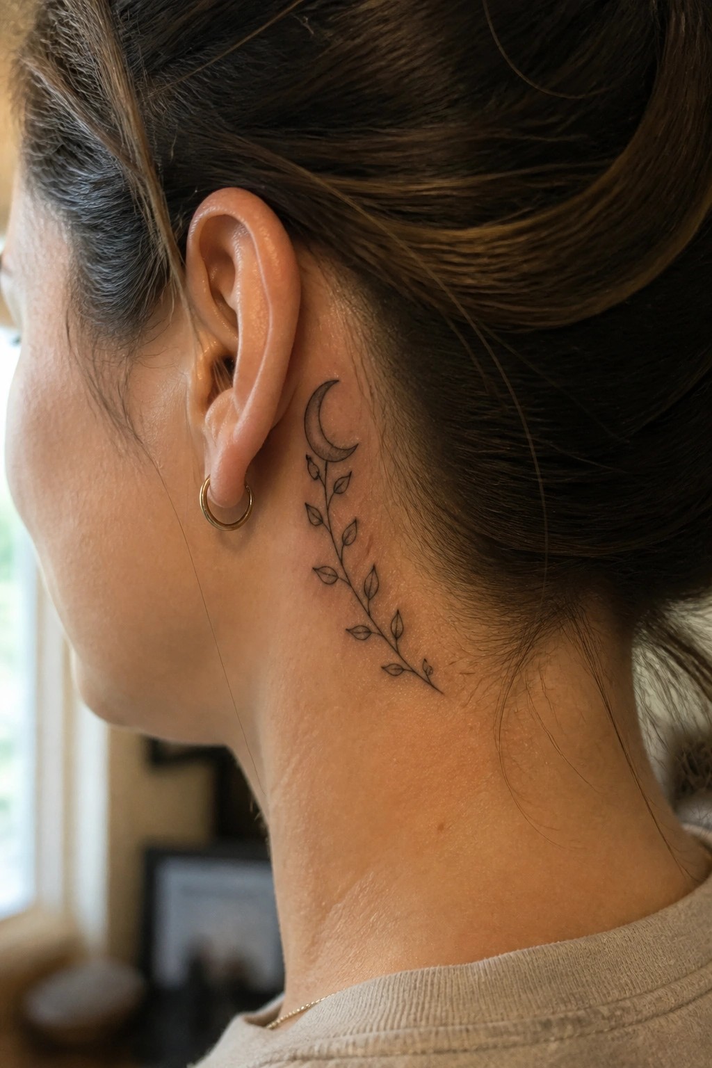 Woman's neck shows crescent moon and leafy vine tattoo behind gold hoop earring