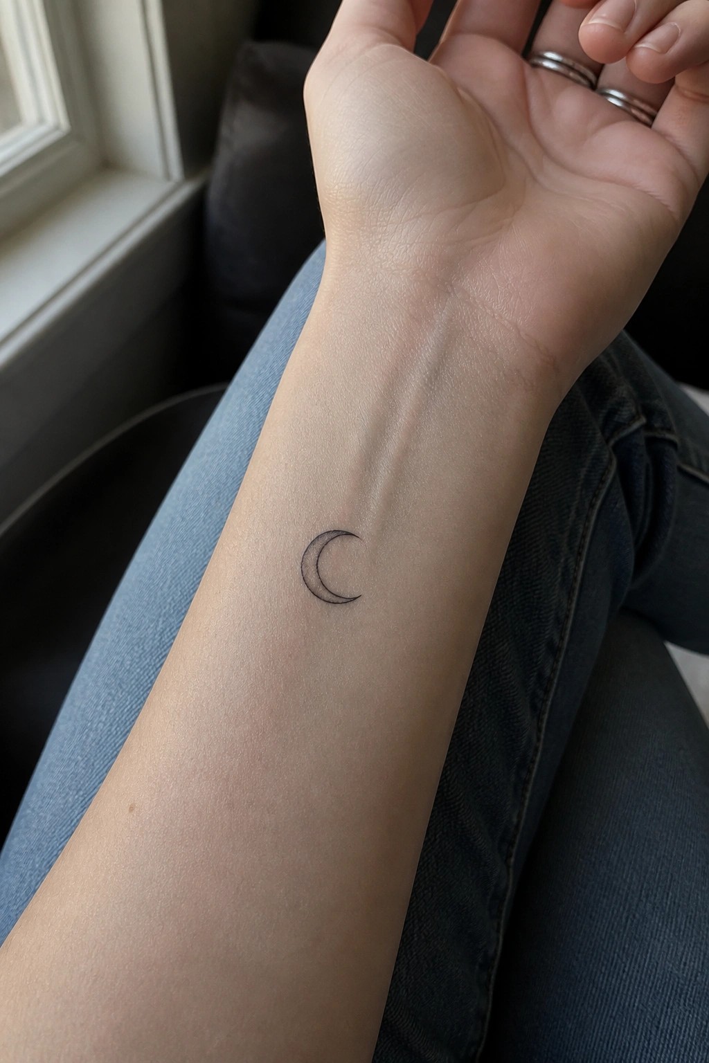 Small crescent moon tattoo on wrist above blue jeans with silver rings