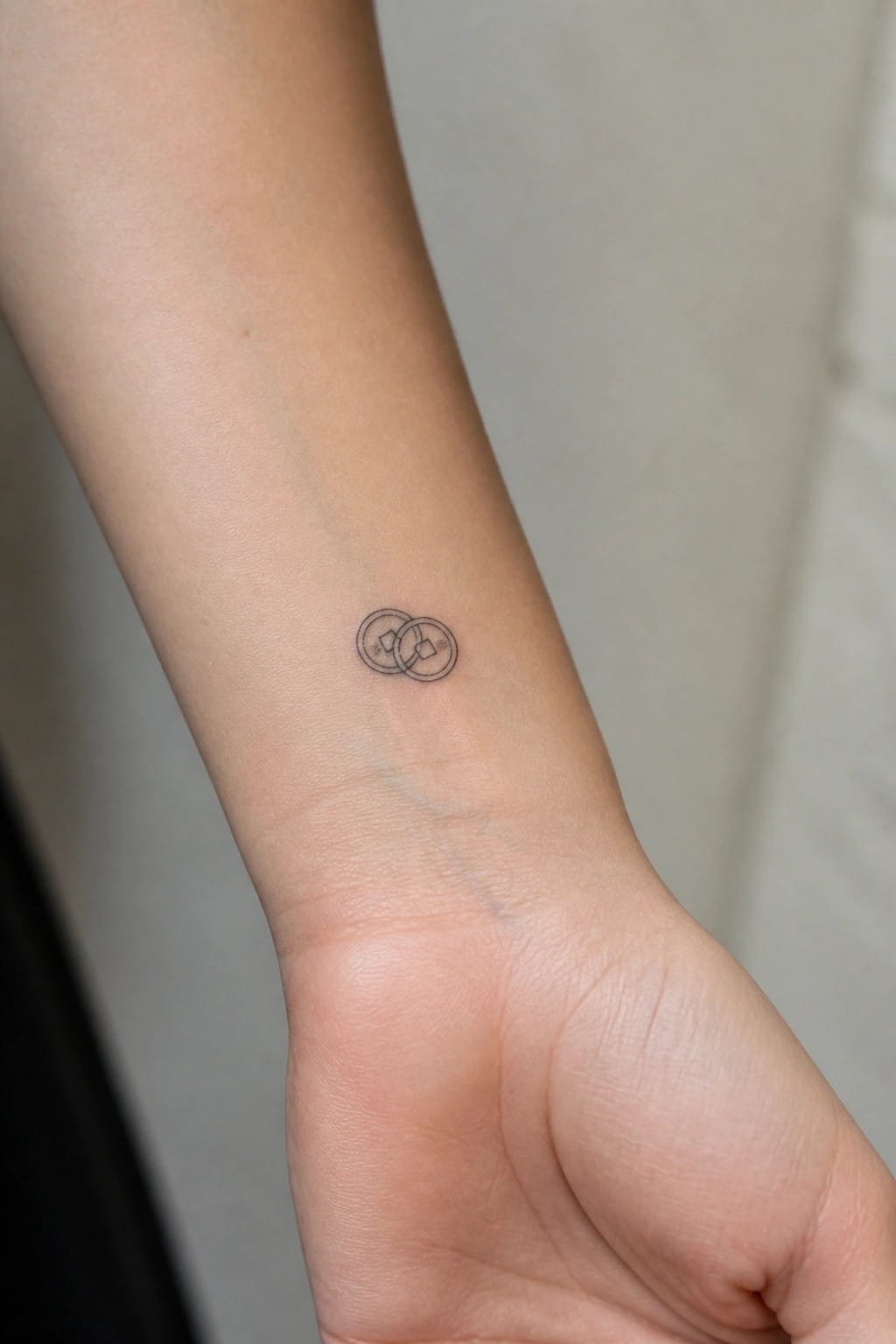Fine line tattoo of two interlocking Chinese coins on the inner wrist.