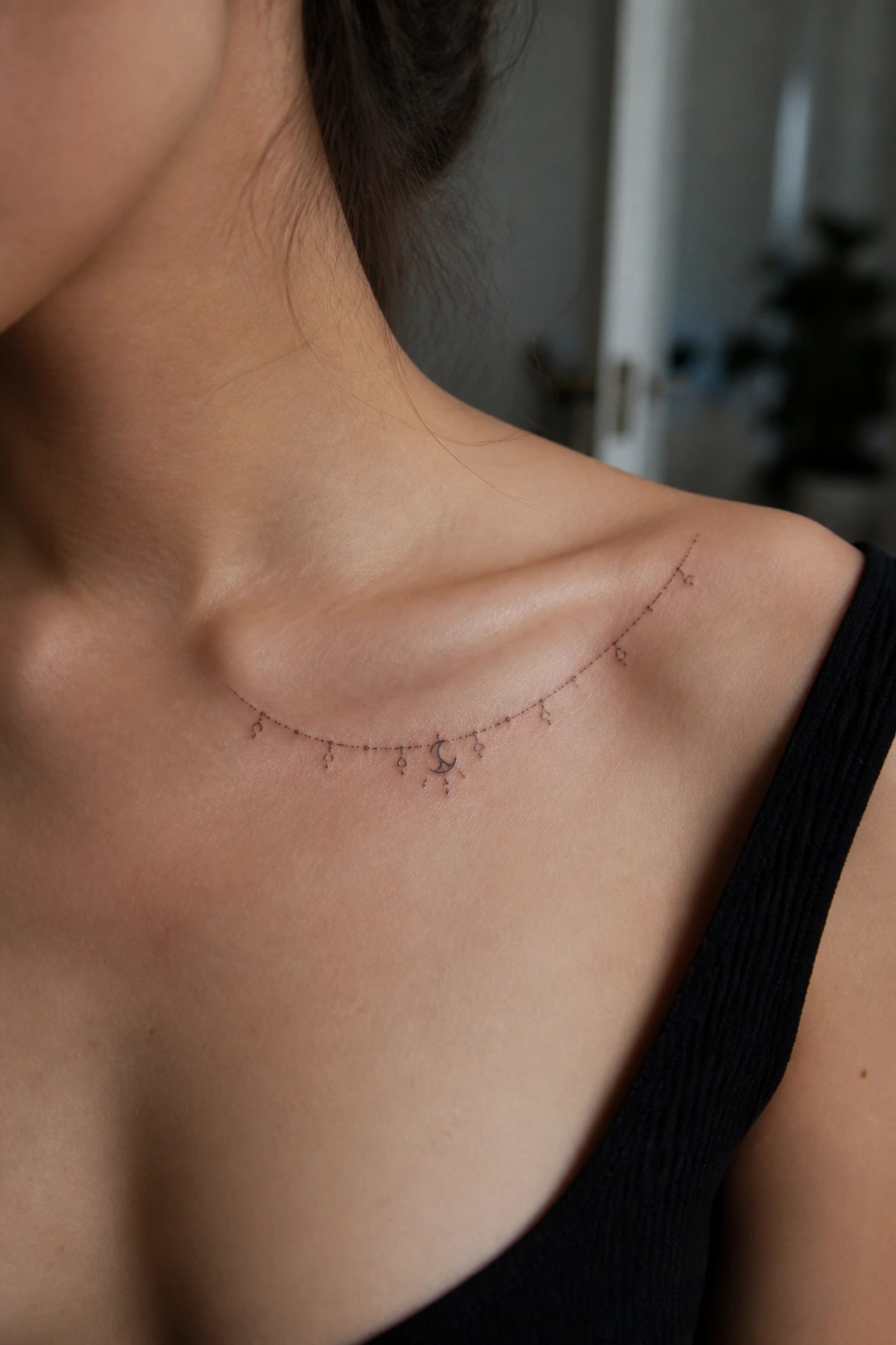 Fine line crescent moon necklace tattoo across a woman's collarbone with small dangling charms and dots
