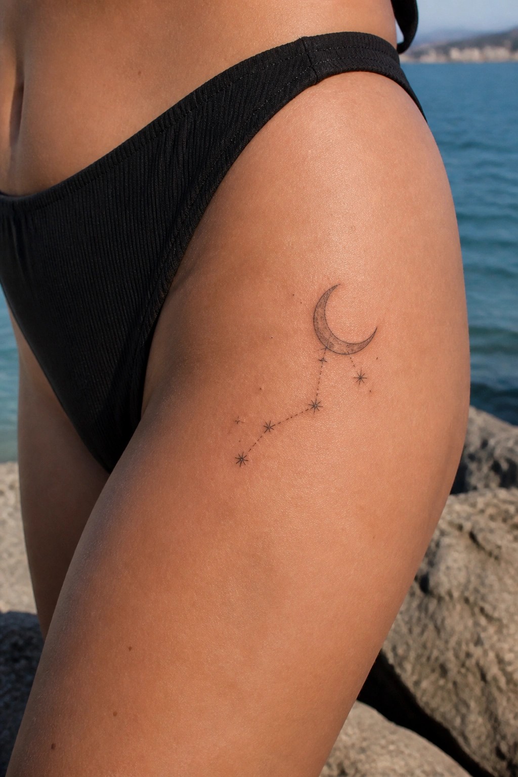 Thigh in black bikini bottoms with crescent moon and star constellation tattoo by the sea.