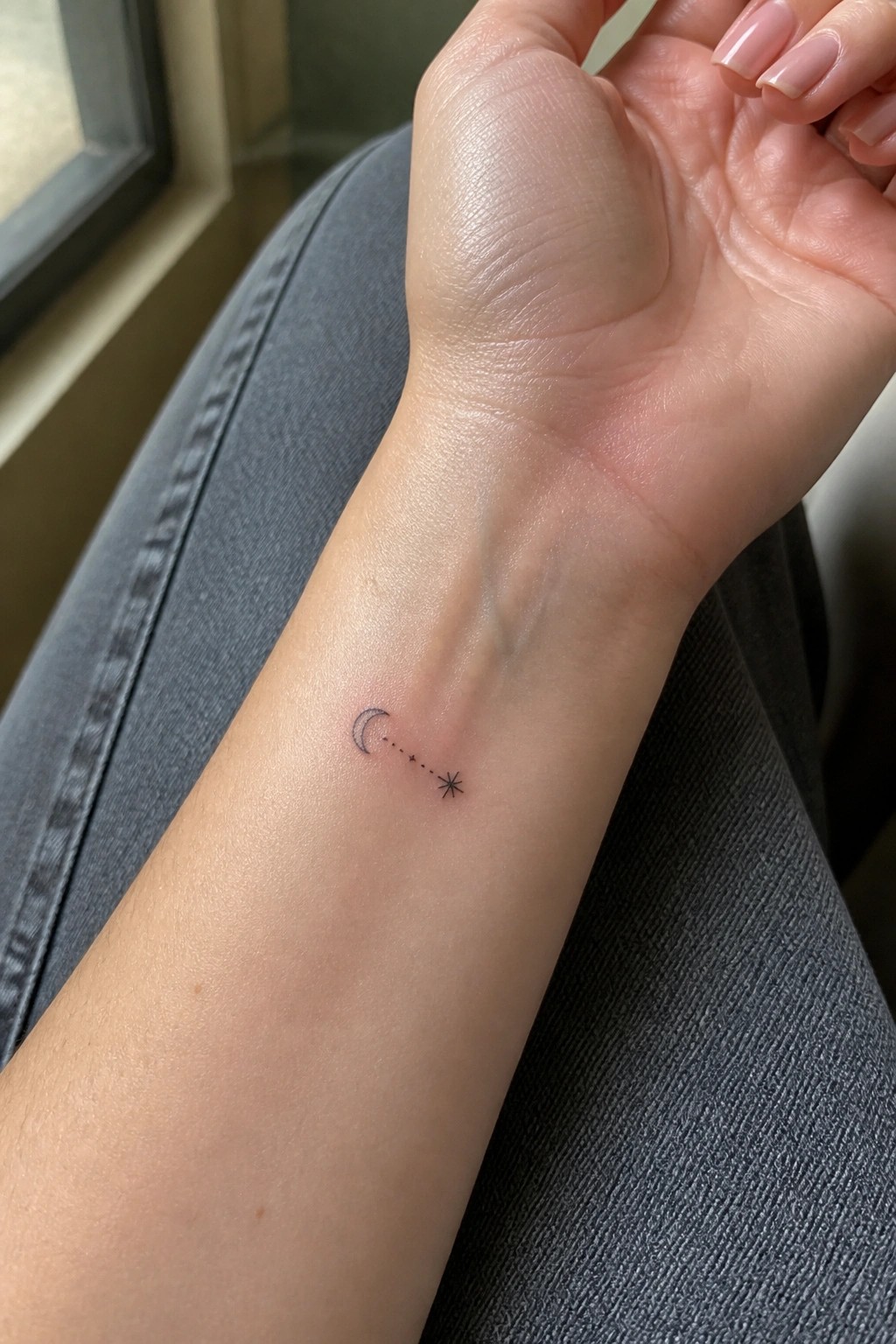 Small crescent moon and dotted star tattoo on wrist over gray denim jeans