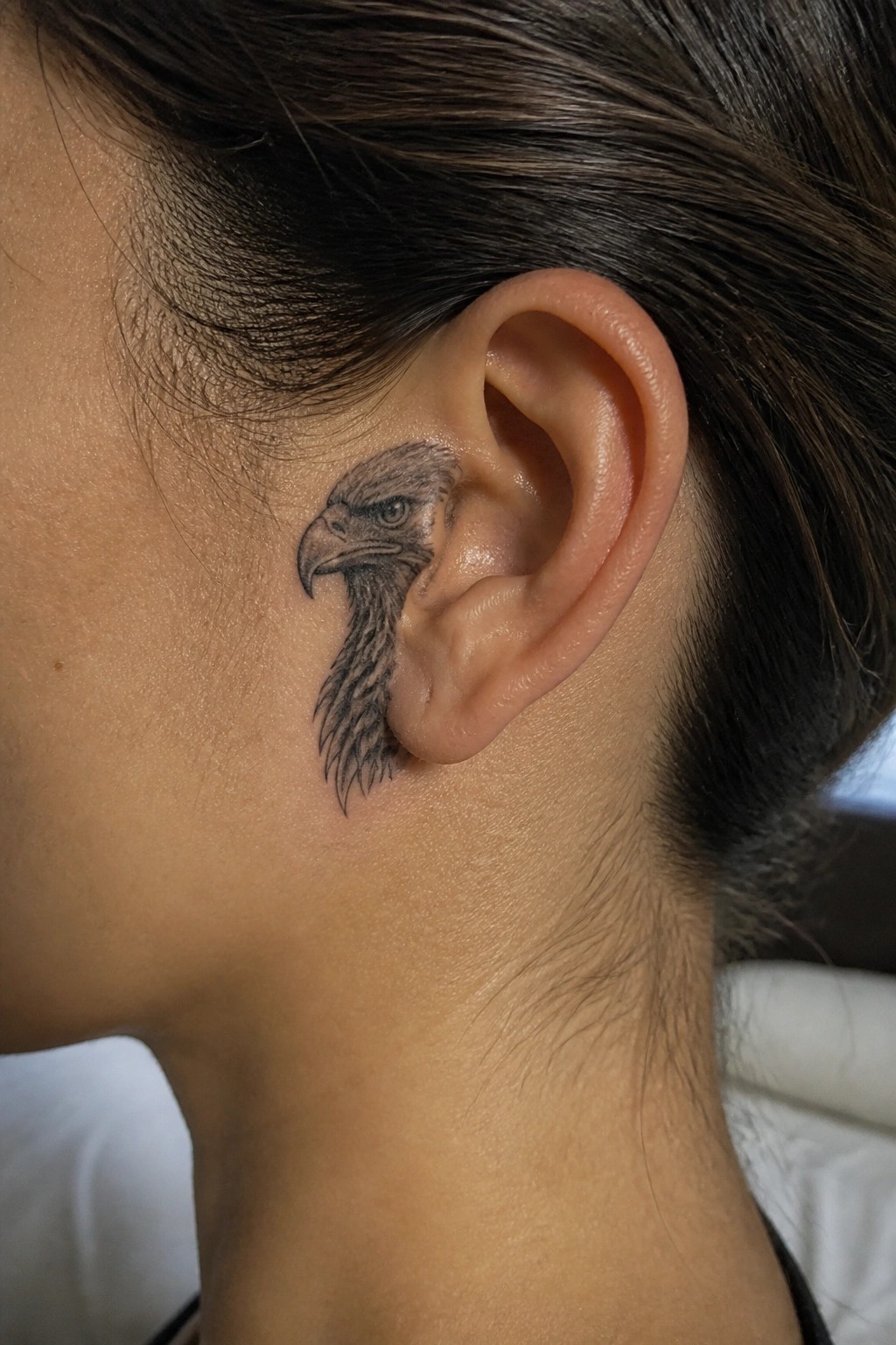 Black and gray eagle head tattoo behind a person's ear on light skin