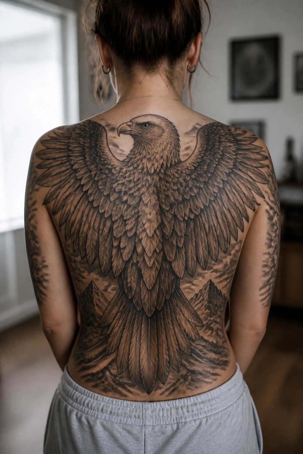 Woman's back with large detailed eagle tattoo, wings spread over shoulders and mountains.