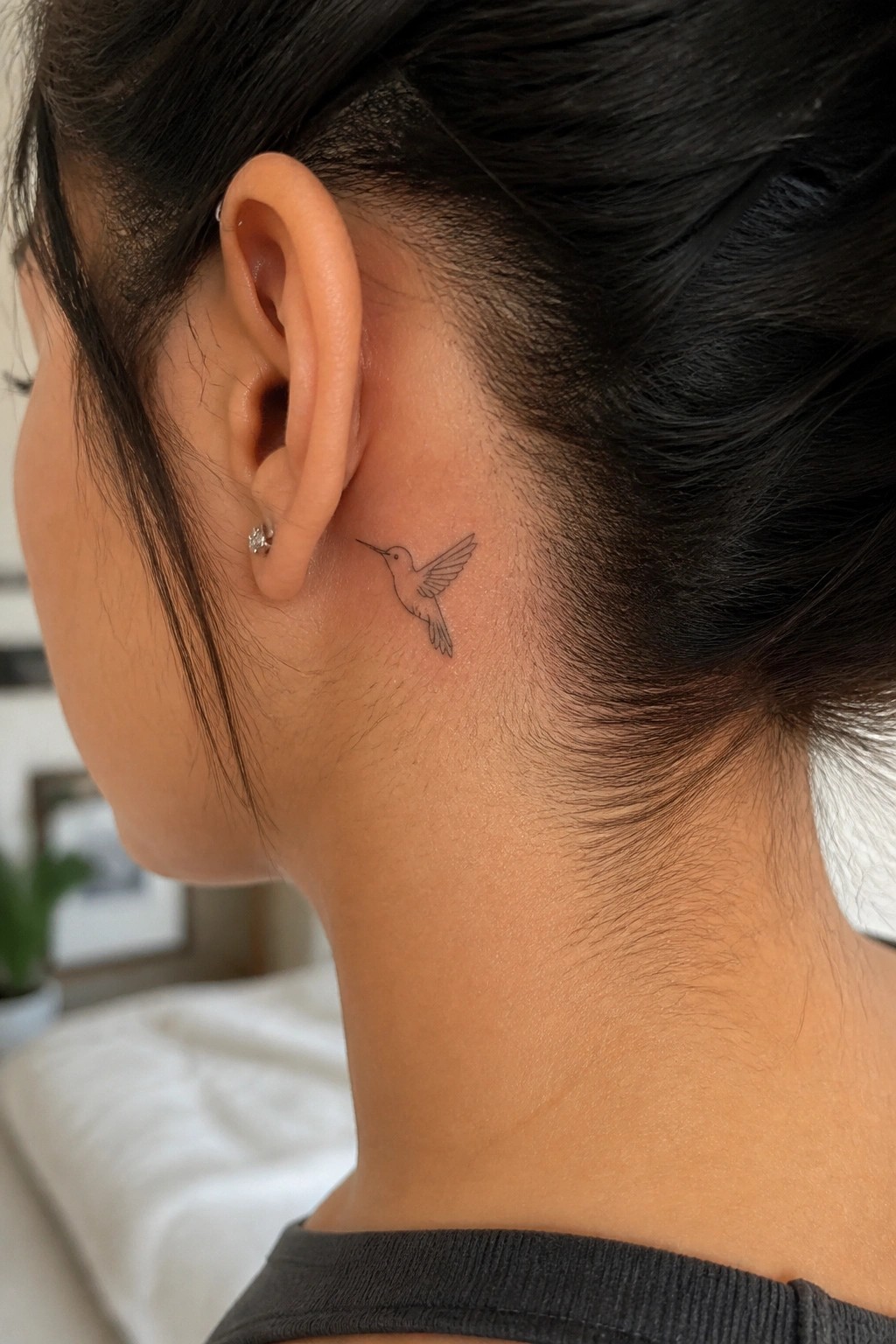 Woman's neck with hummingbird tattoo behind ear, dark bun, and stud earring