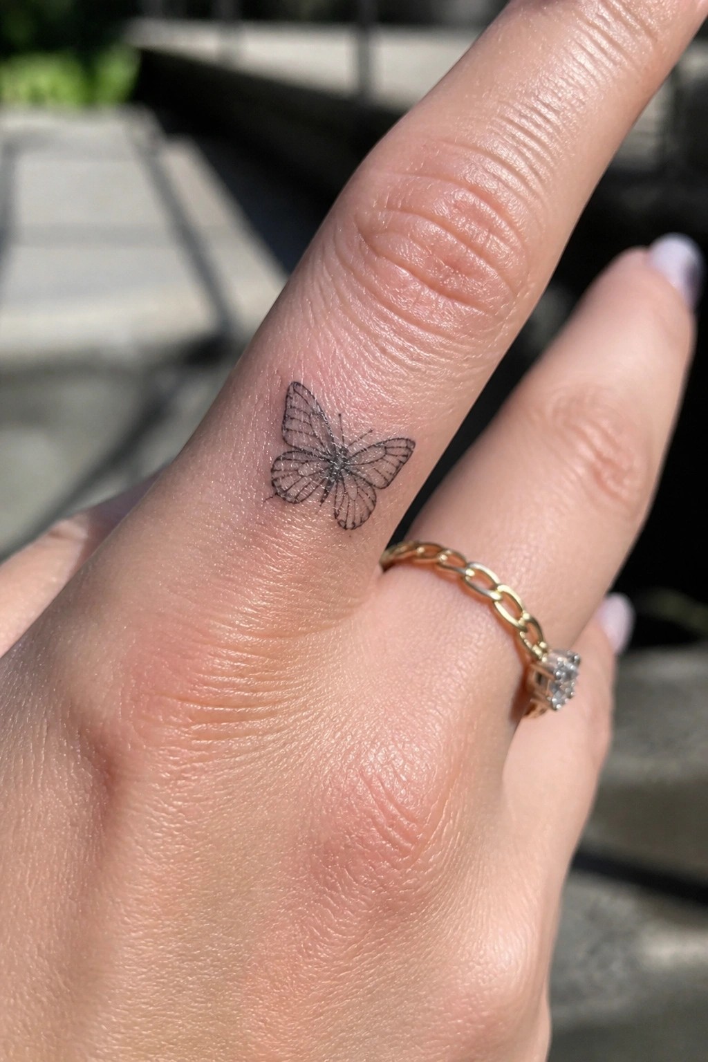 Small fine line butterfly tattoo placed on the side of a finger beside a gold ring.