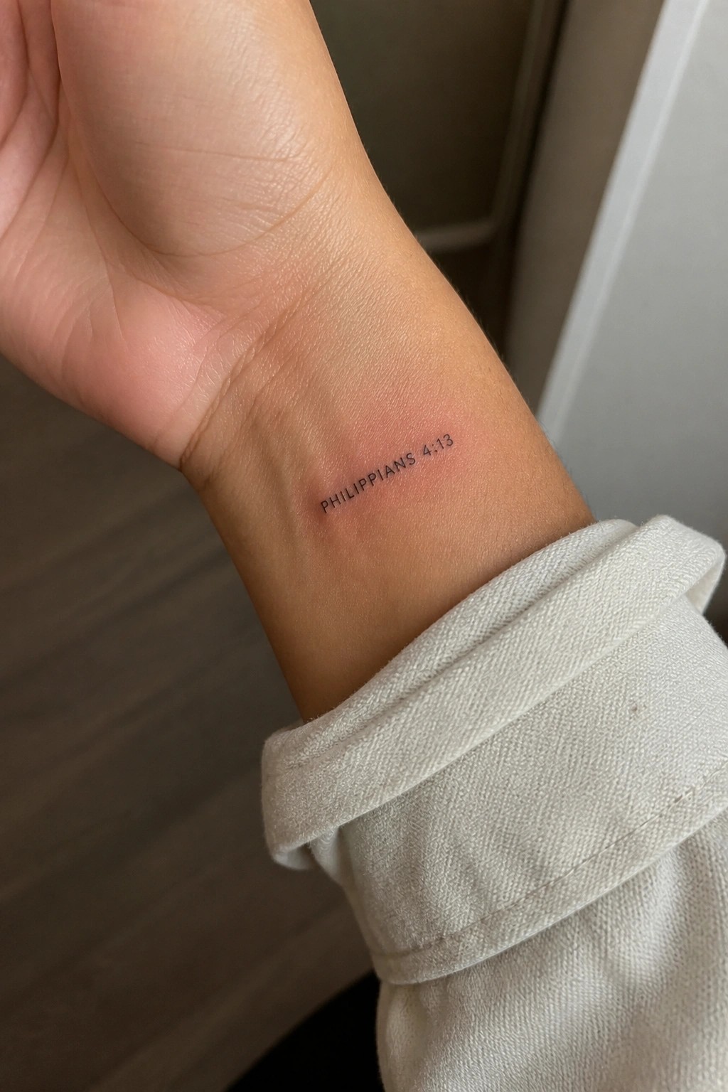 Wrist with Philippians 4:13 tattoo on light skin beside beige fabric cuff
