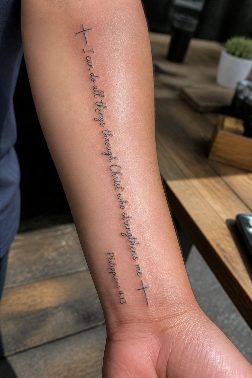 Cursive black tattoo of Philippians 4:13 on forearm near blue shirt and table