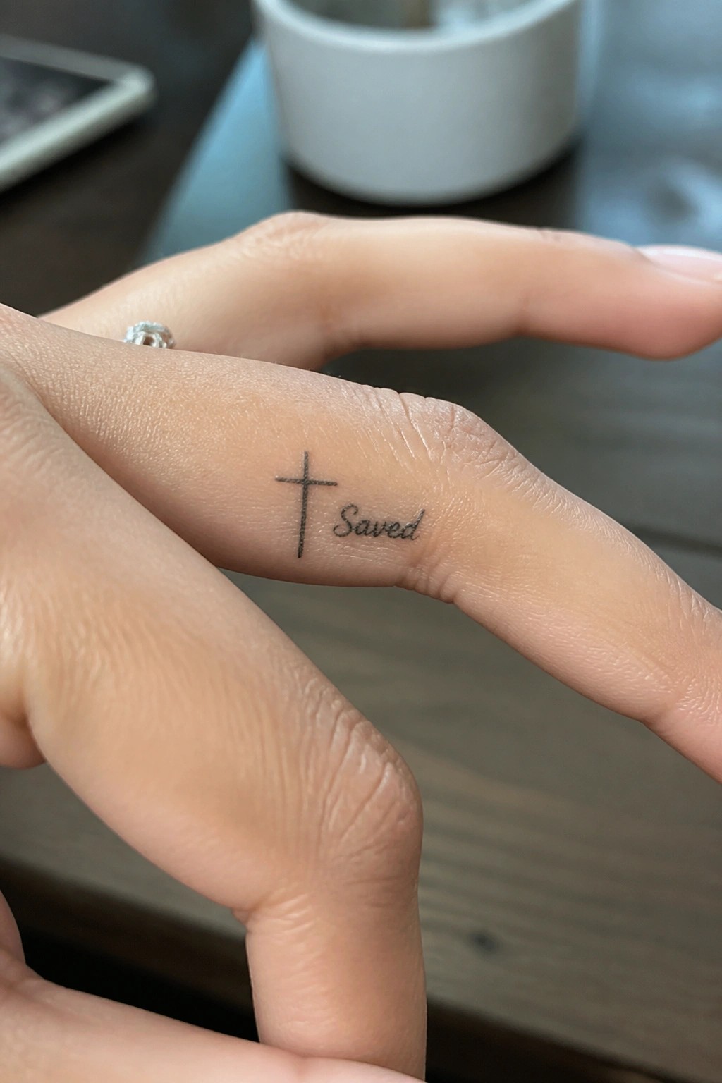 Small fine line cross tattoo with the word saved on the side of a finger