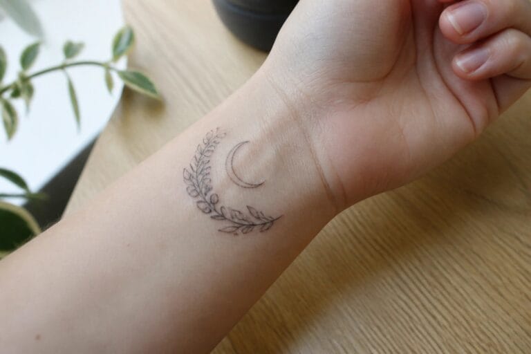 Minimalist crescent moon tattoo with leafy branches on light-skinned wrist over wood