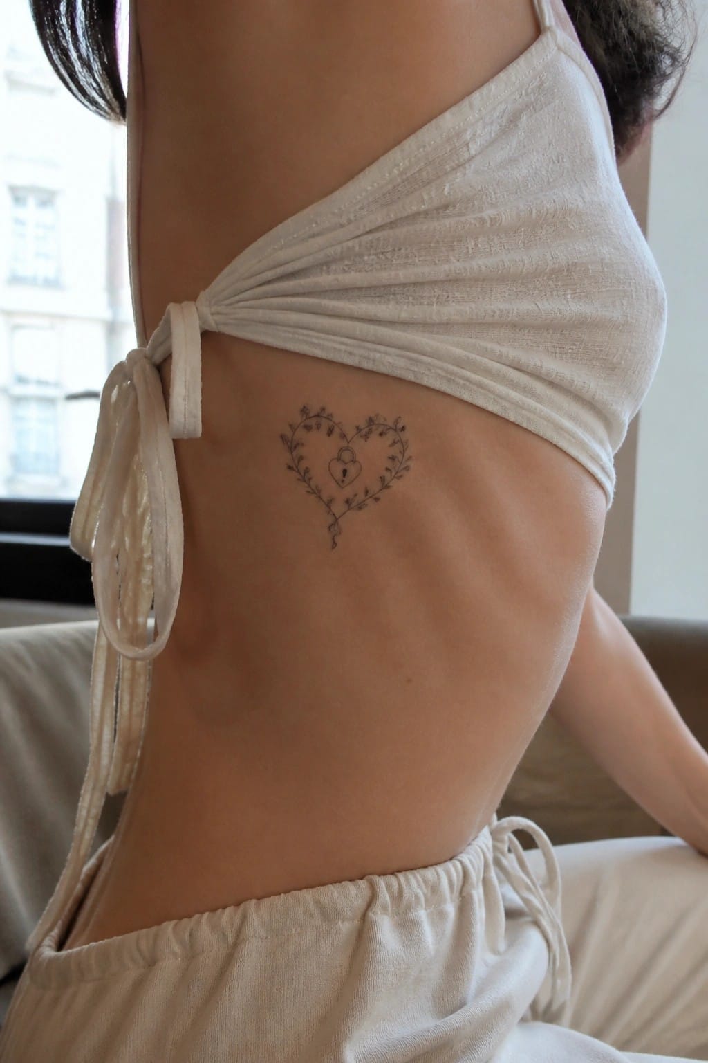White halter top tied at side reveals vine heart tattoo with padlock on ribs.