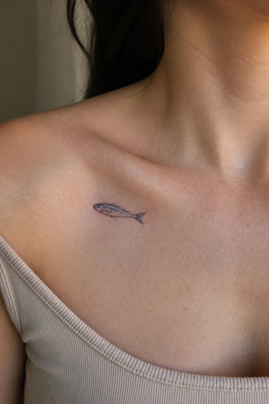 Small blue fish tattoo on shoulder above beige ribbed tank top strap