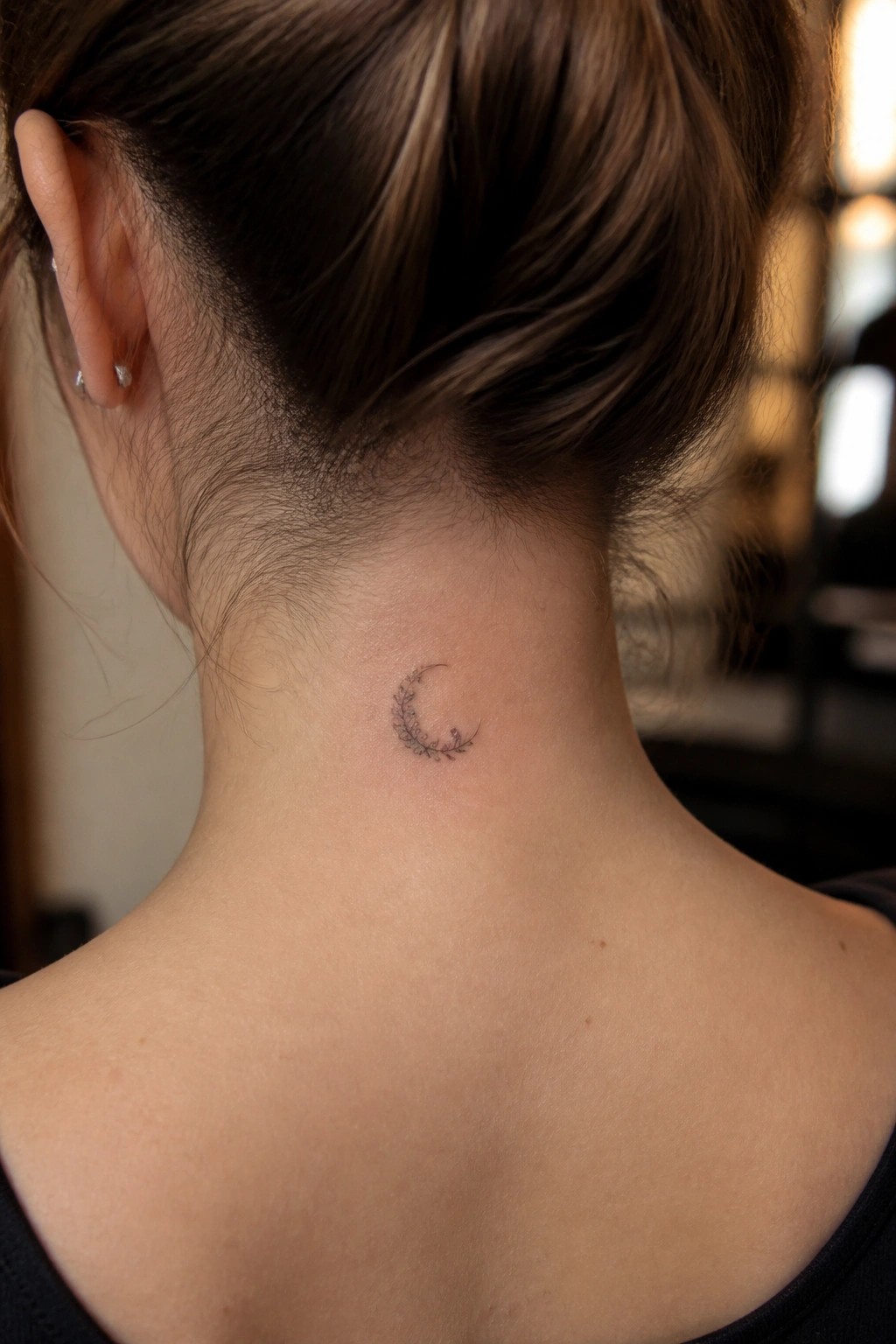 Crescent moon floral tattoo on woman's nape, brown hair in bun, black top