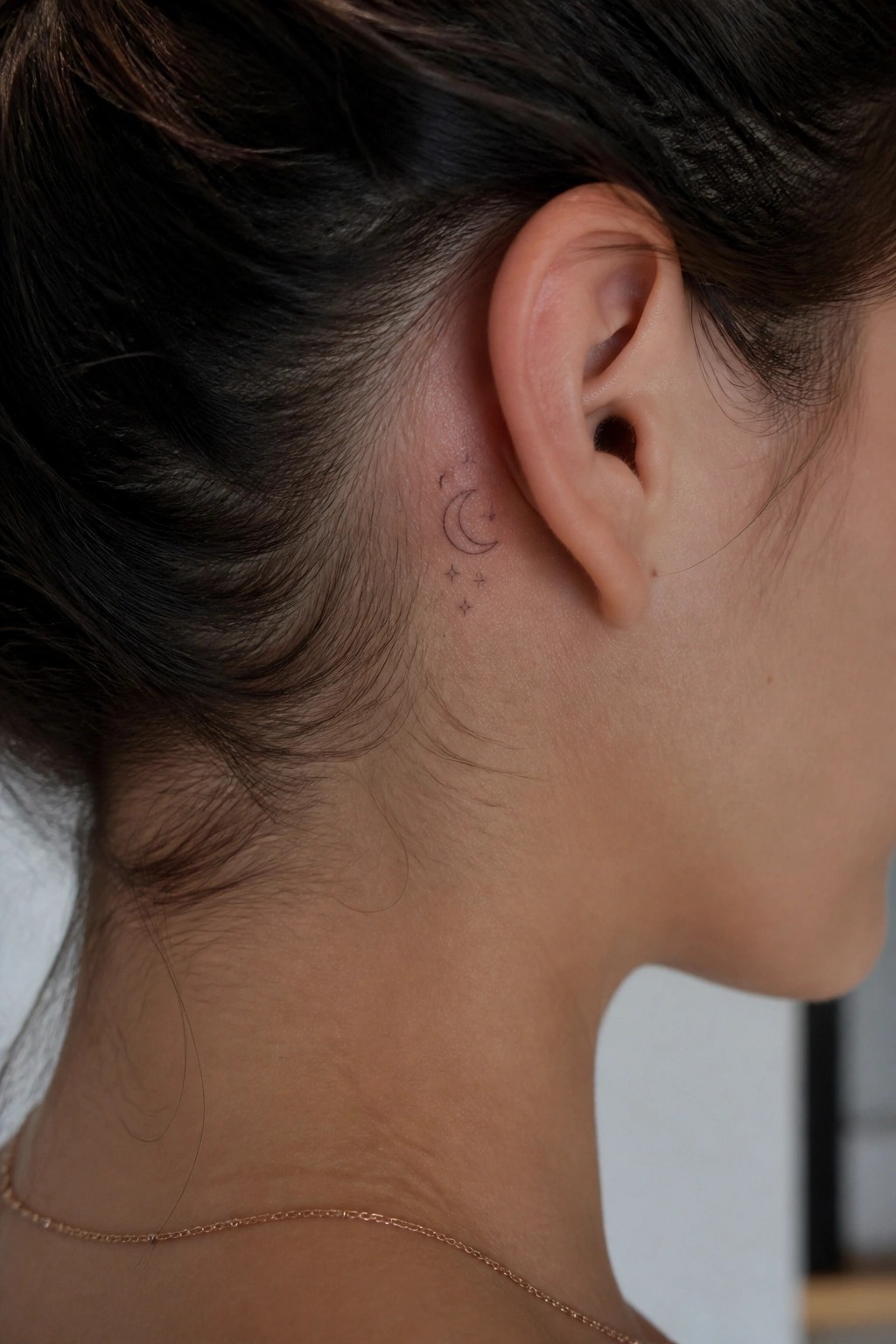 Woman's neck with crescent moon and stars tattoo behind ear, thin gold necklace.