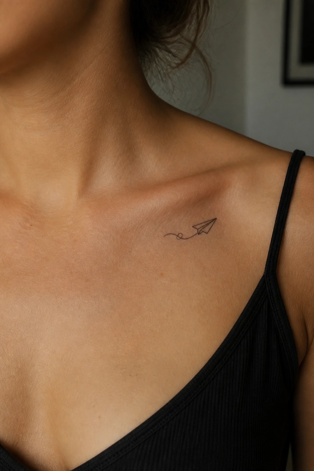 Paper airplane tattoo on woman's collarbone above black tank top strap.