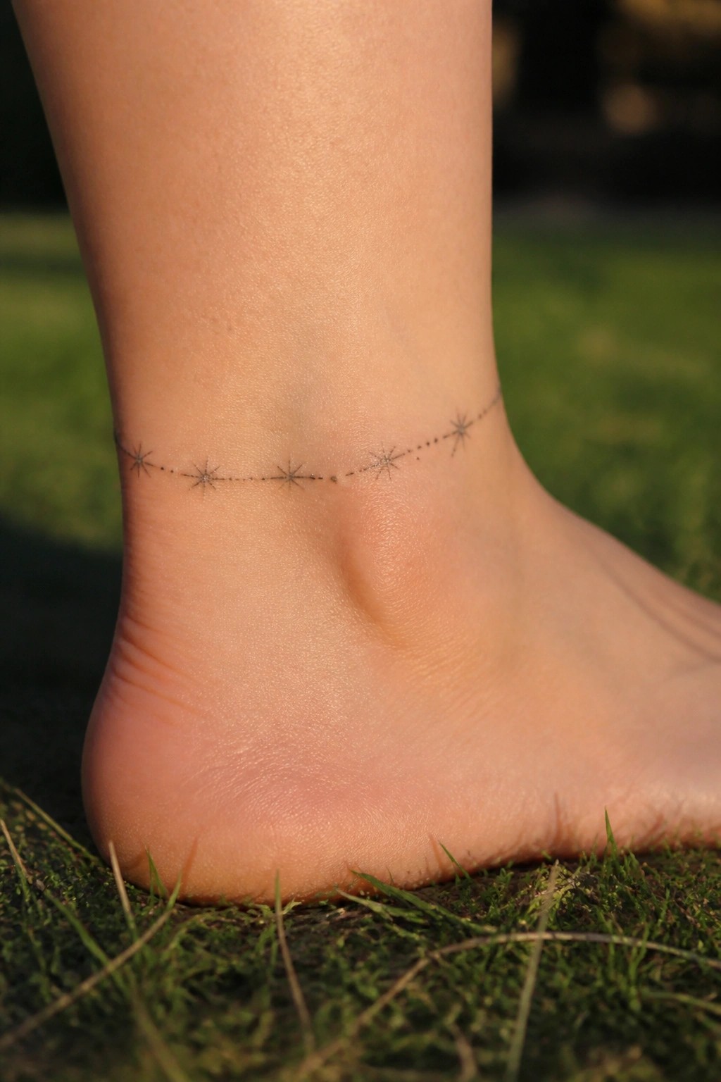 Fine line tattoo of small stars connected by dots wrapping around an ankle on grass
