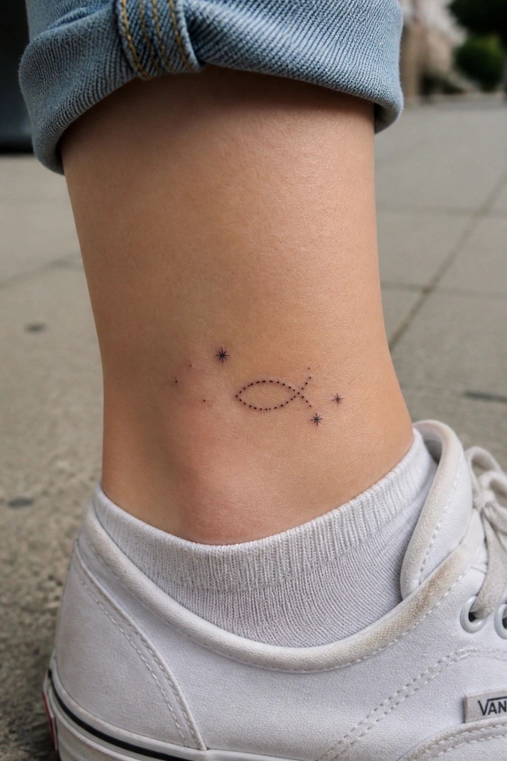 Dotted fish tattoo with stars on ankle below cuffed blue jeans and white sneaker