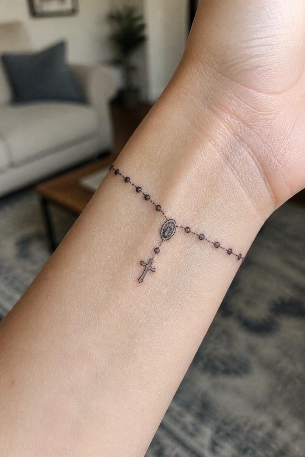 Fine line black rosary tattoo on the inner wrist with beaded chain, oval medallion, and small cross.