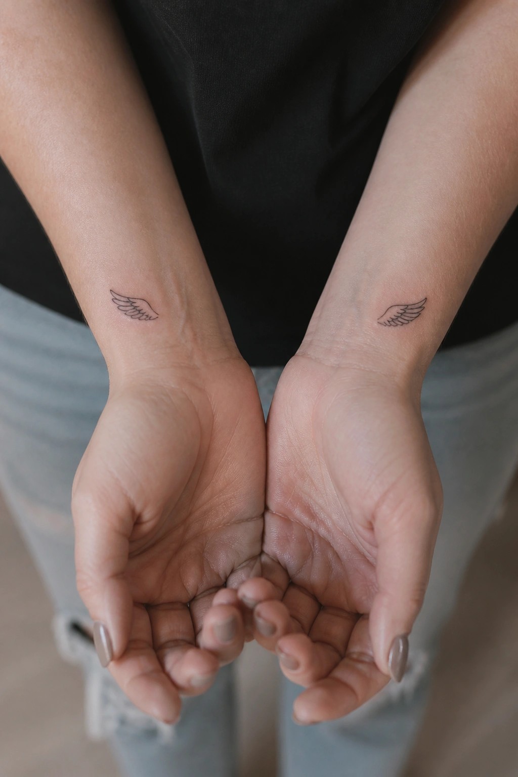 Palms-up hands with matching wing tattoos on wrists, black shirt and ripped jeans.