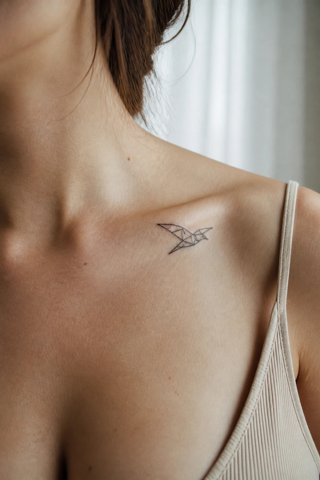 Geometric bird tattoo on fair-skinned shoulder beside beige tank top strap