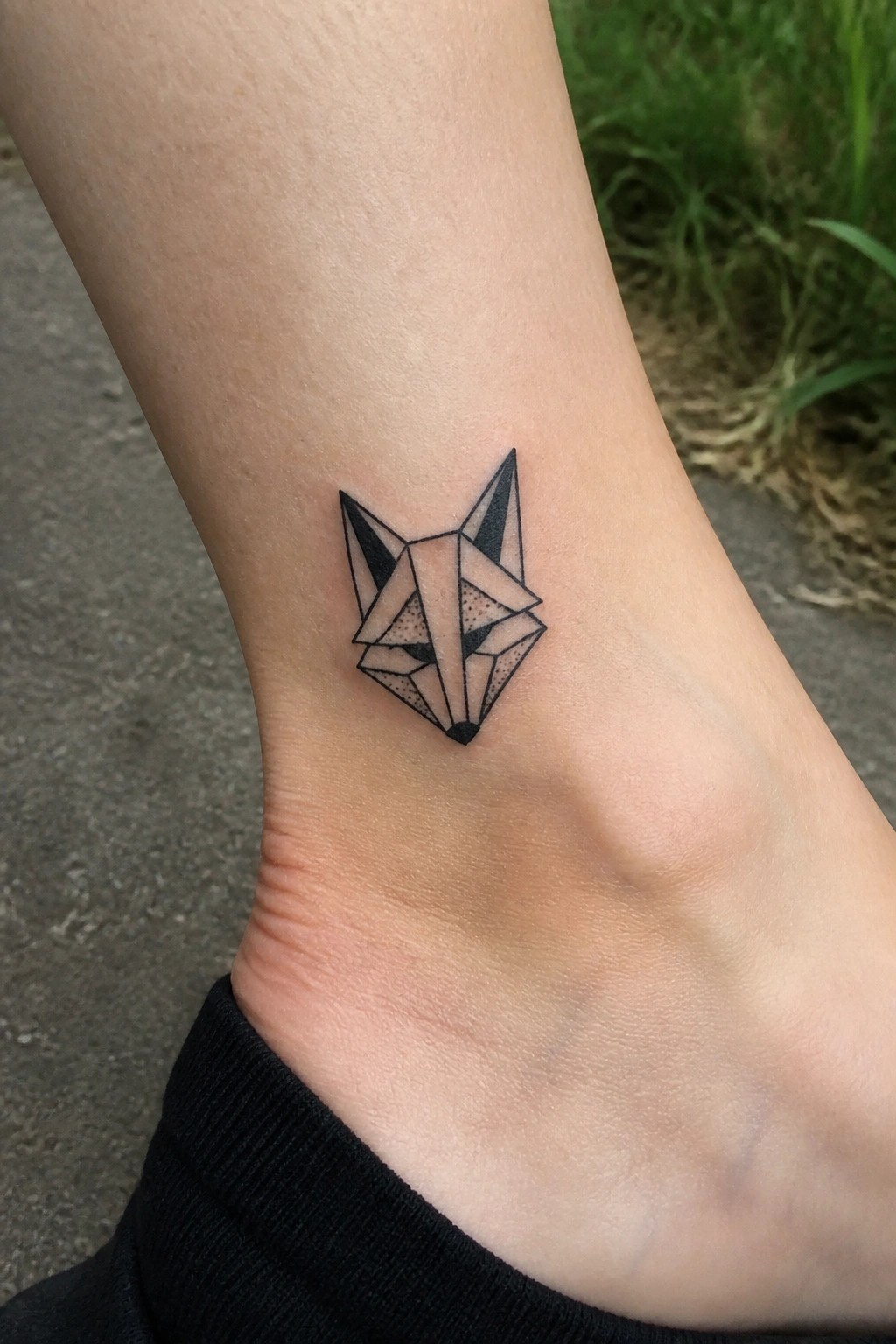 Small geometric fox head tattoo on the ankle in black linework with polygonal shapes and dotted shading.