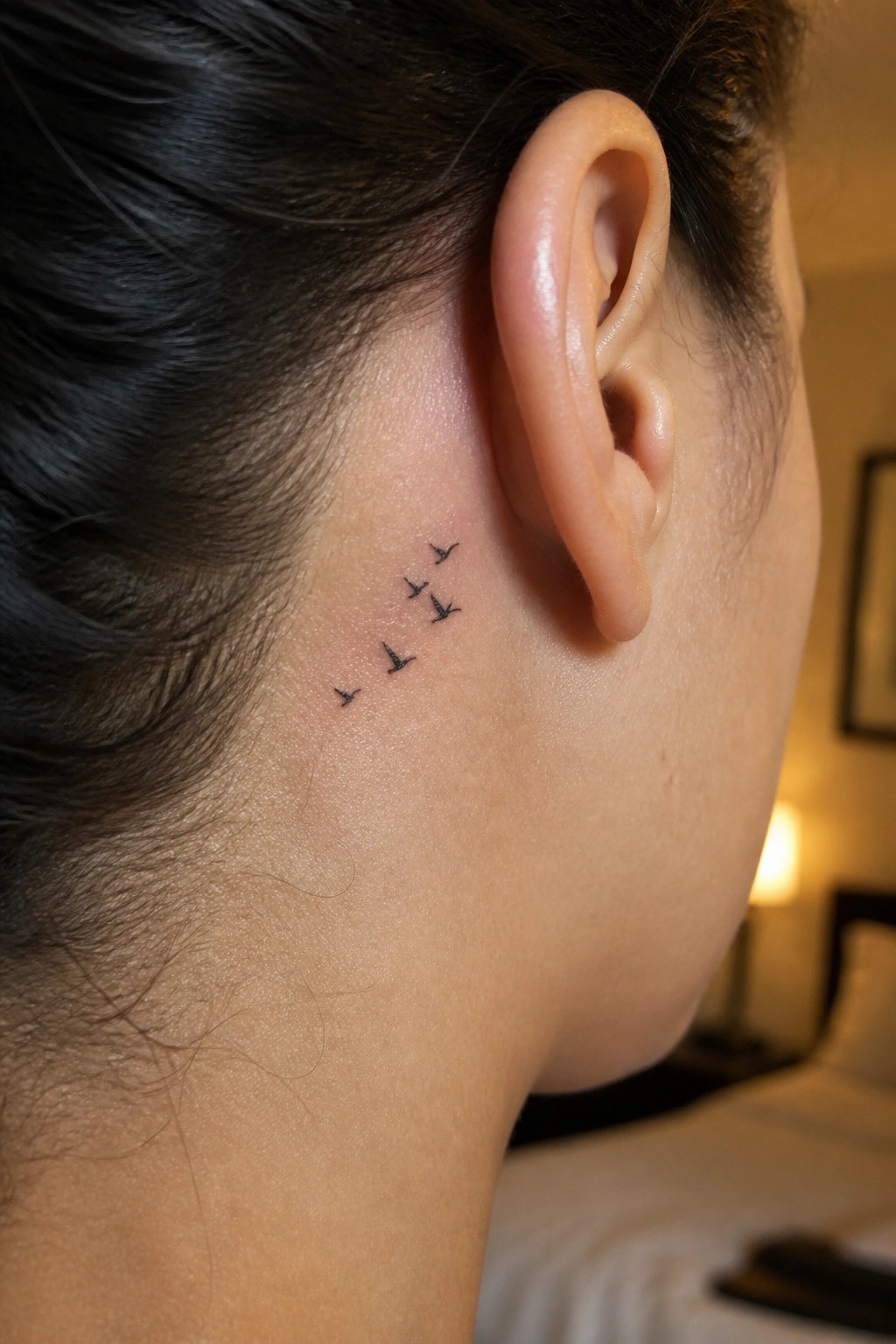 Small black bird tattoos in flight behind a person's ear with dark hair