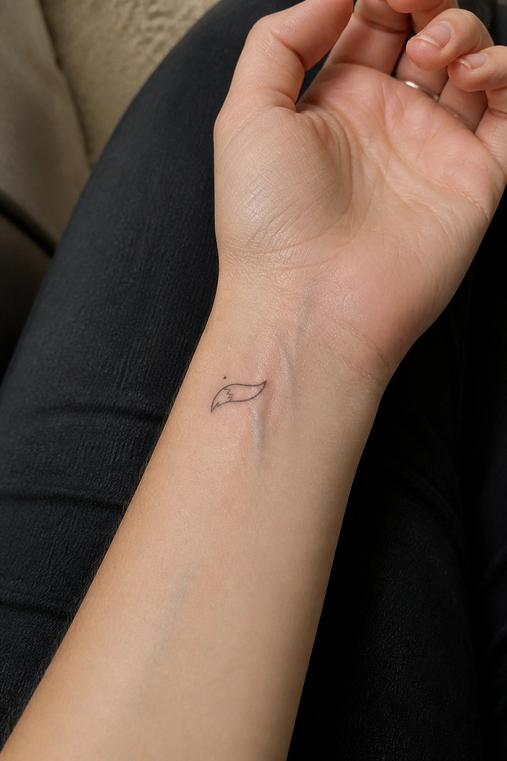 Light-skinned wrist with small black wing tattoo resting on dark clothing