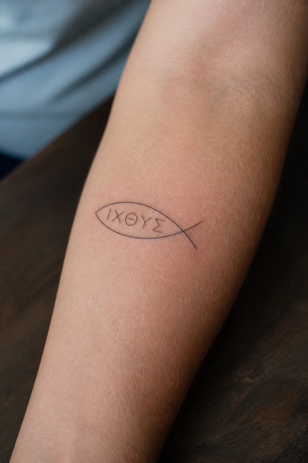 Minimalist line tattoo of the Christian fish symbol with Greek letters inside, placed on the inner forearm.