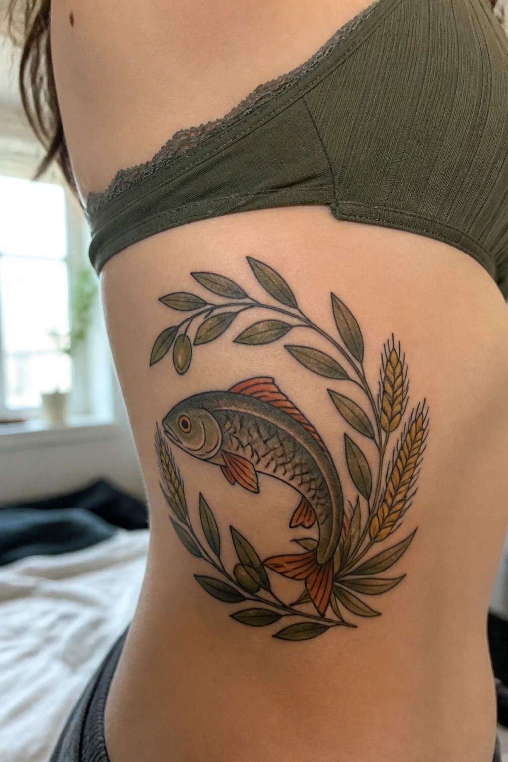 A fish tattoo on the ribs surrounded by olive branches and wheat in a circular wreath style.