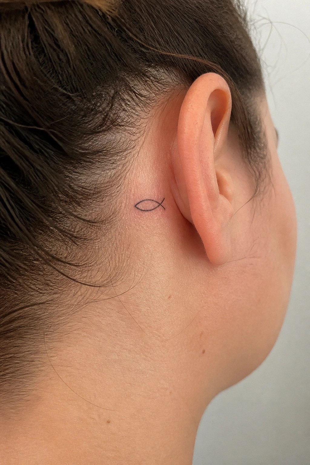 Tiny fish tattoo behind the ear on light skin with dark pulled-back hair