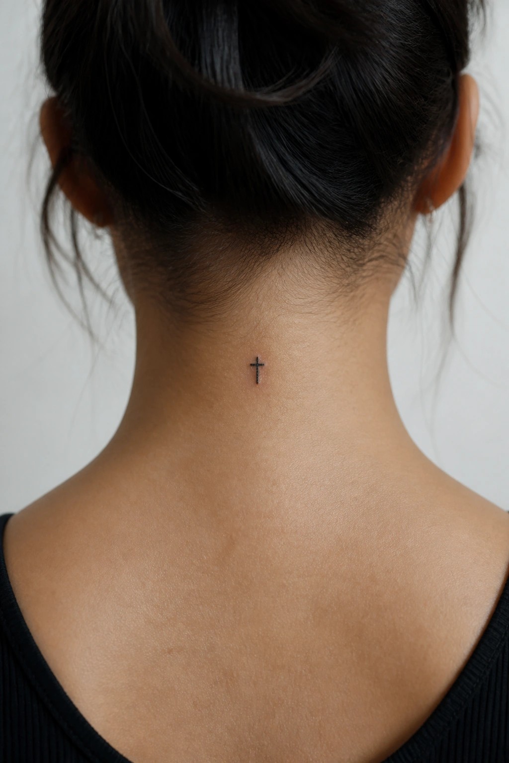 Small black cross tattoo on woman's neck below dark bun, black top.