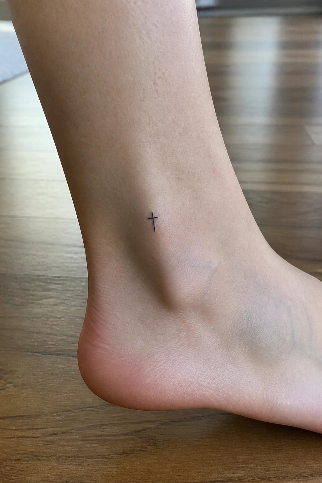 Bare foot ankle with tiny black cross tattoo above heel on wood floor