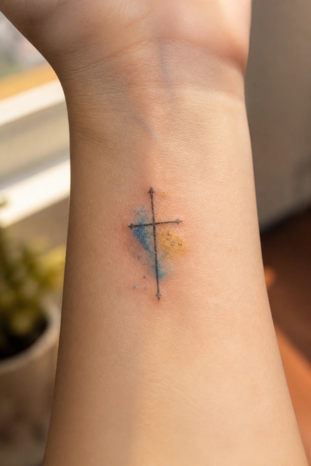 Small black cross tattoo with blue and yellow watercolor splashes on wrist