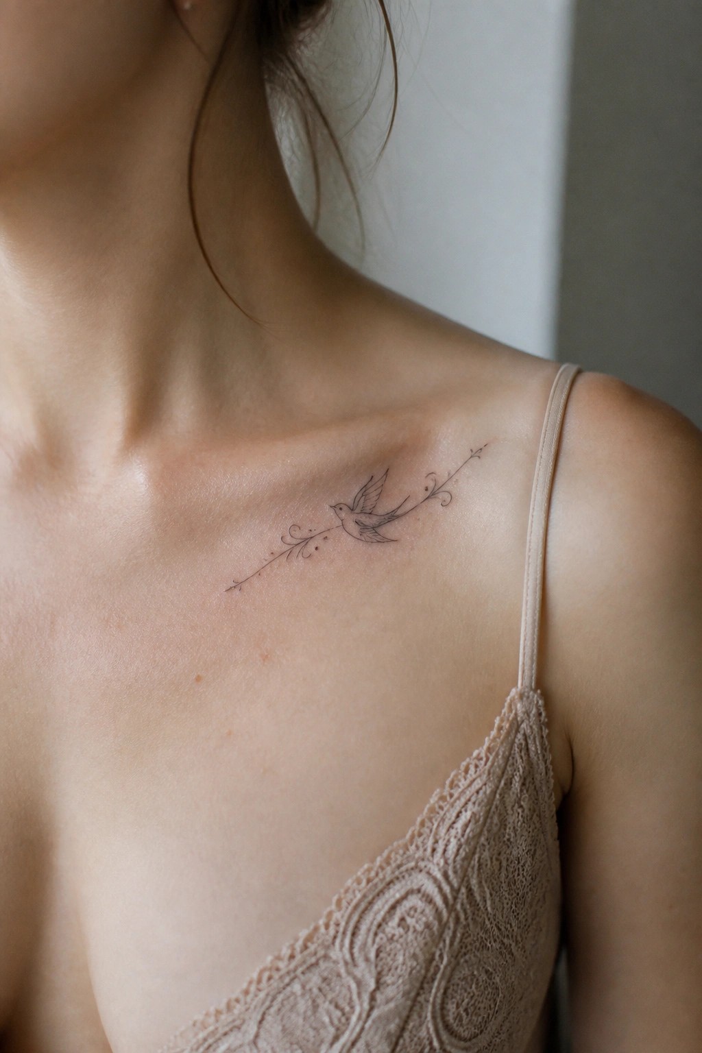 Woman's shoulder with delicate bird tattoo above beige lace camisole strap