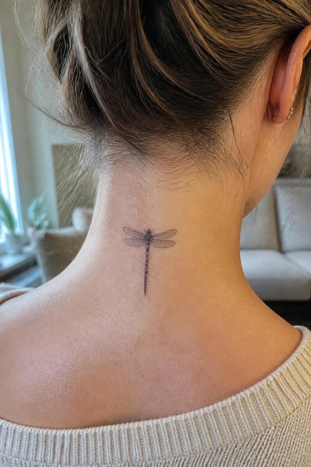 Small dragonfly tattoo on nape of neck below hair bun in cream sweater