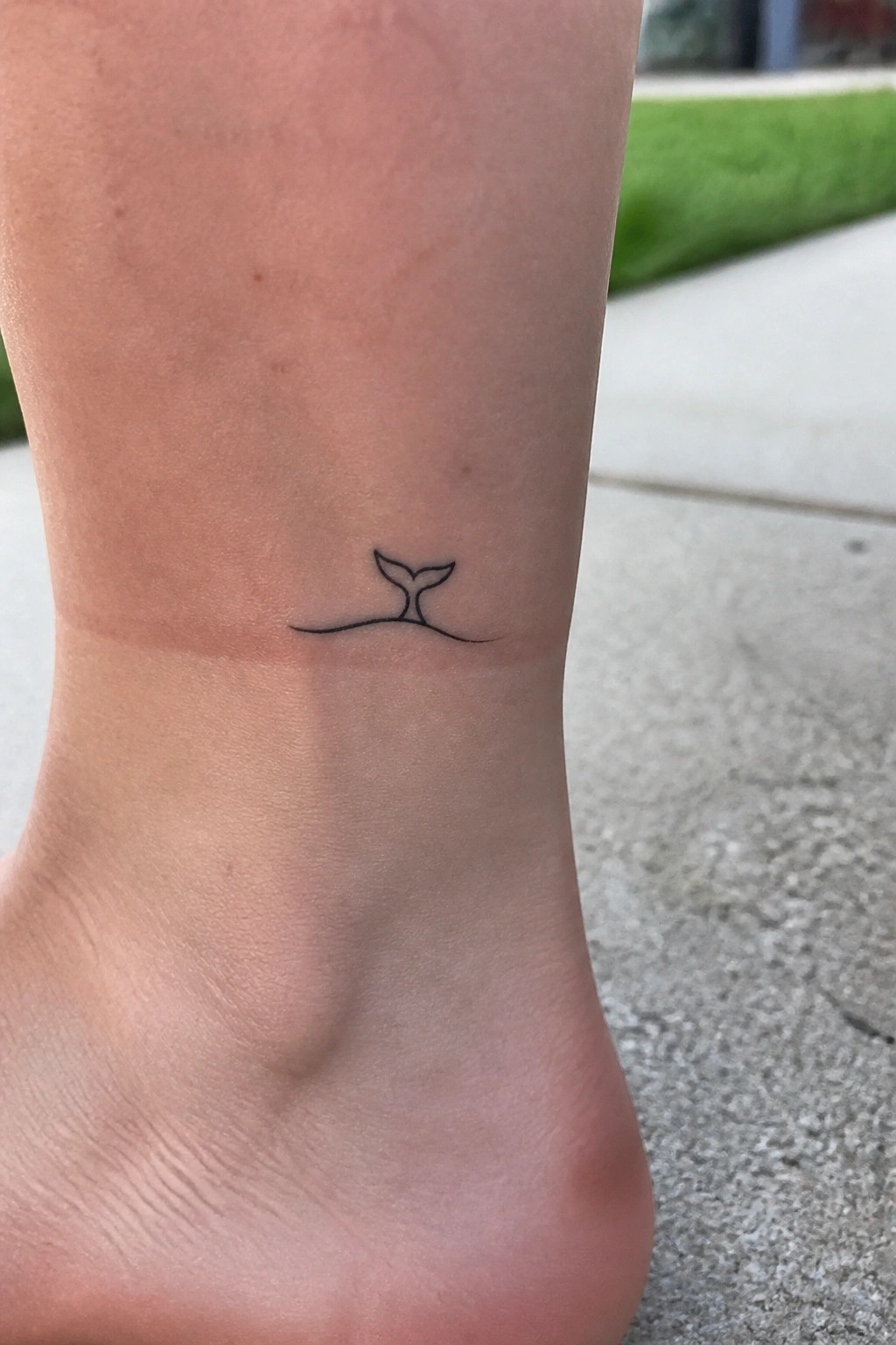 Small black whale tail tattoo with wave line on bare ankle