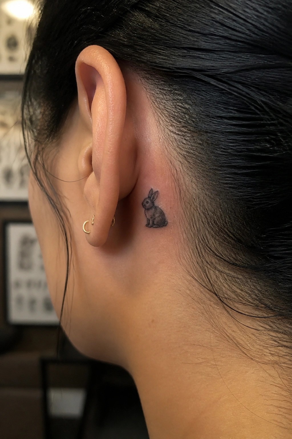 Small black rabbit tattoo behind ear with gold crescent moon earrings.