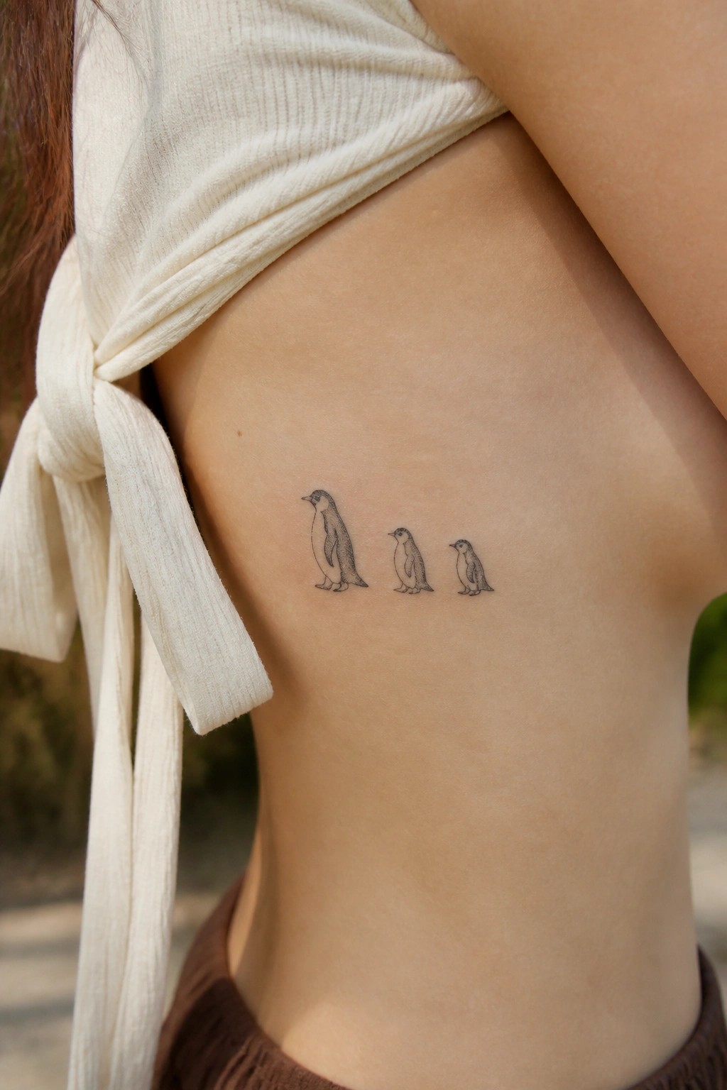 Three decreasing-size penguin tattoos on skin under white knotted top.