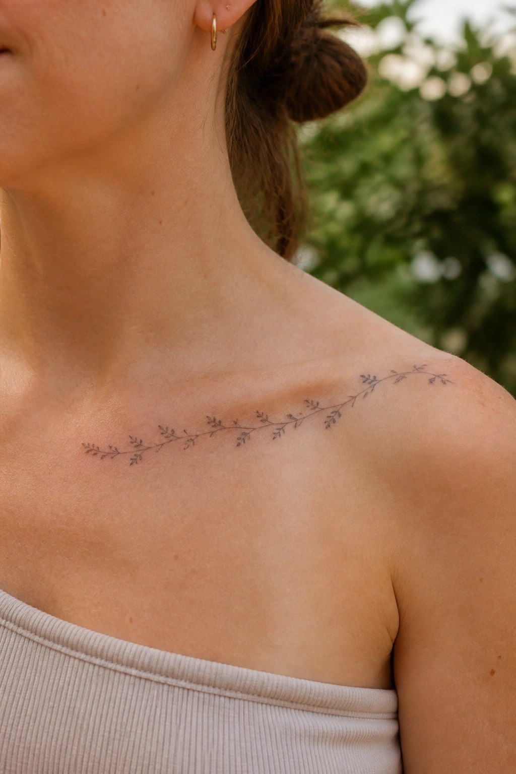 Woman's collarbone with delicate leafy branch tattoo, beige ribbed top, gold earring