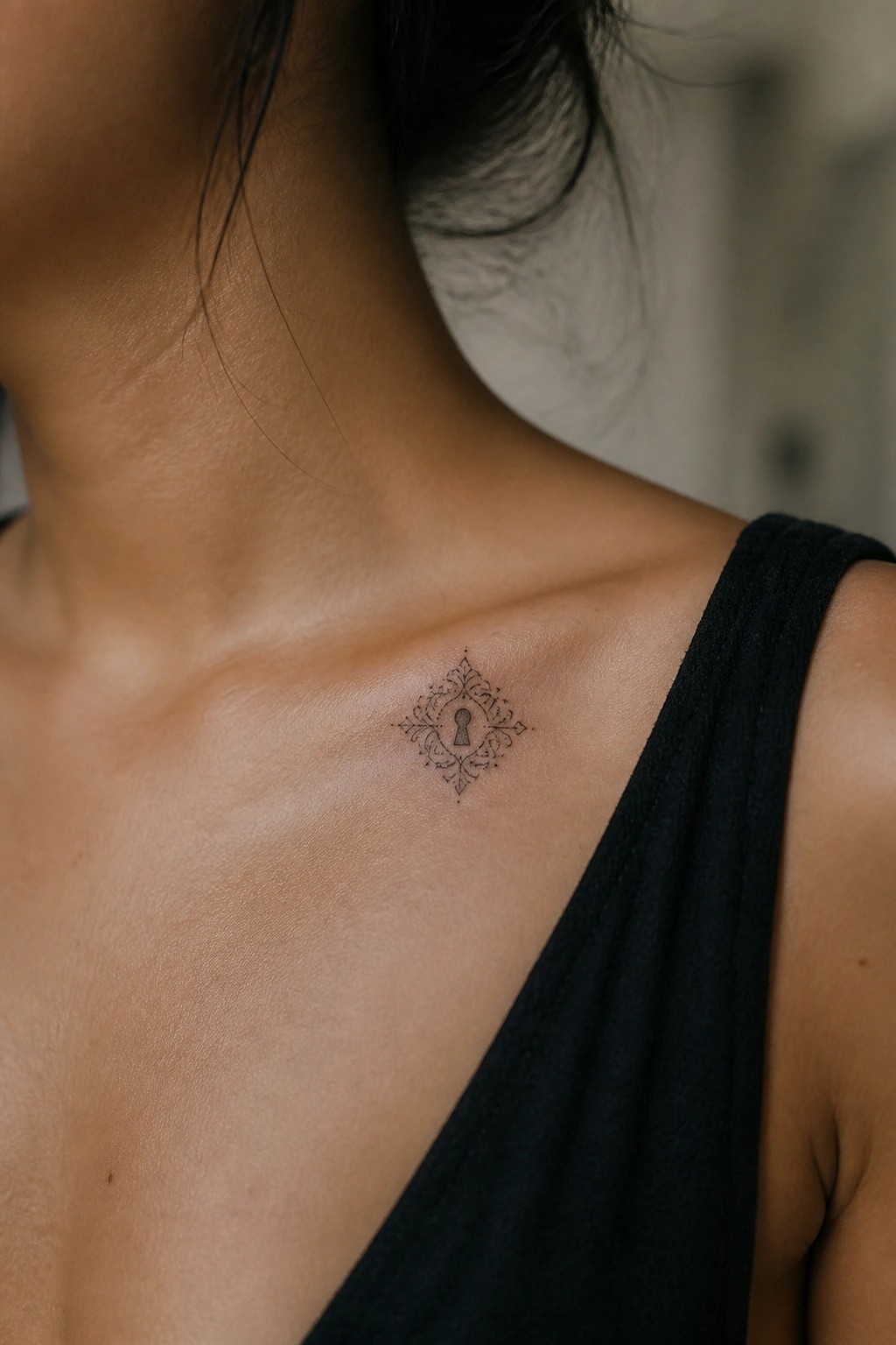 Ornate keyhole tattoo on collarbone above black top, dark hair framing tanned skin.