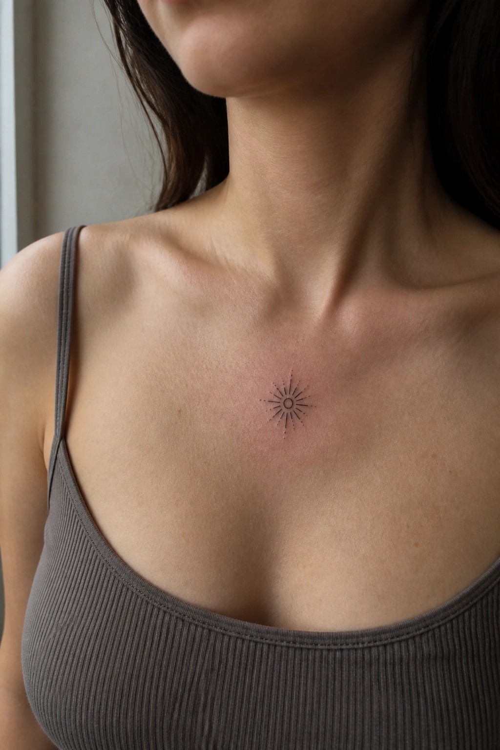 Delicate sun tattoo on reddened chest above gray ribbed tank top neckline